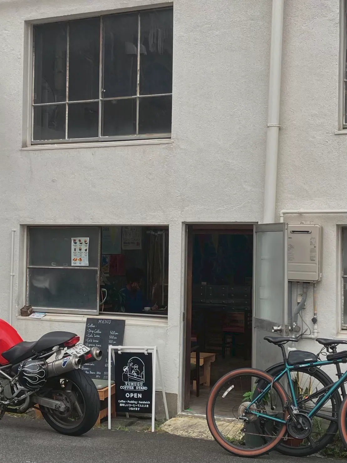 TUMUGU COFFEE STANDの外観。白い建物の一角にあり、メニューを書いた黒板とOPENの看板、窓越しに店内が見える入口